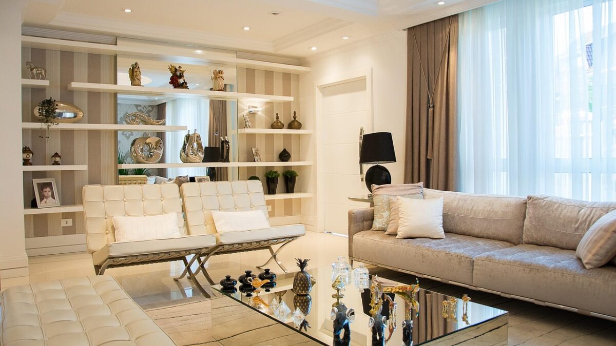 Salon moderne aux tons crème et beige avec canapé en tissu, deux fauteuils en cuir capitonné, table basse en verre miroir, étagères blanches et rideaux taupe.