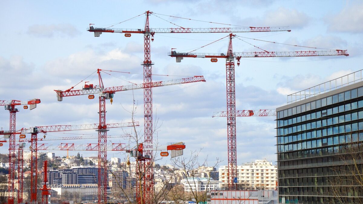 Plusieurs grues de chantier rouges et blanches dominent la scène devant une ville aux immeubles blancs et une tour de bureaux vitrée à droite, sous un ciel nuageux.
