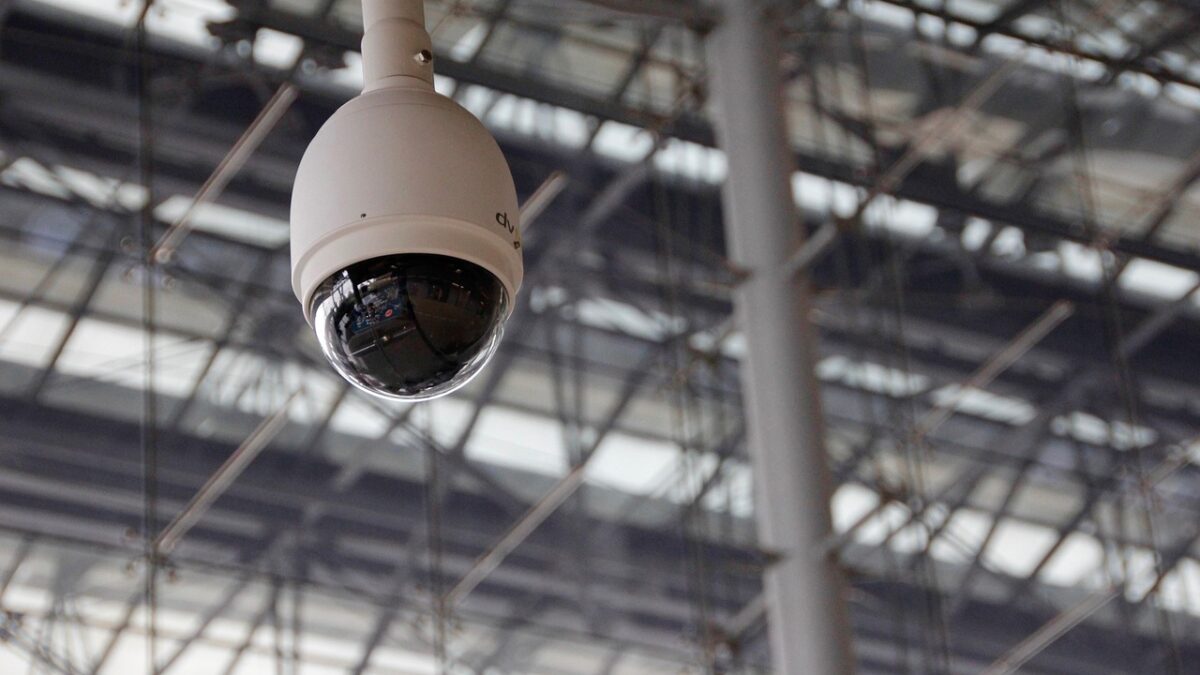 Caméra de surveillance blanche en forme de dôme fixée à un plafond métallique, à côté d'un poteau vertical, sous une lumière diffuse.
