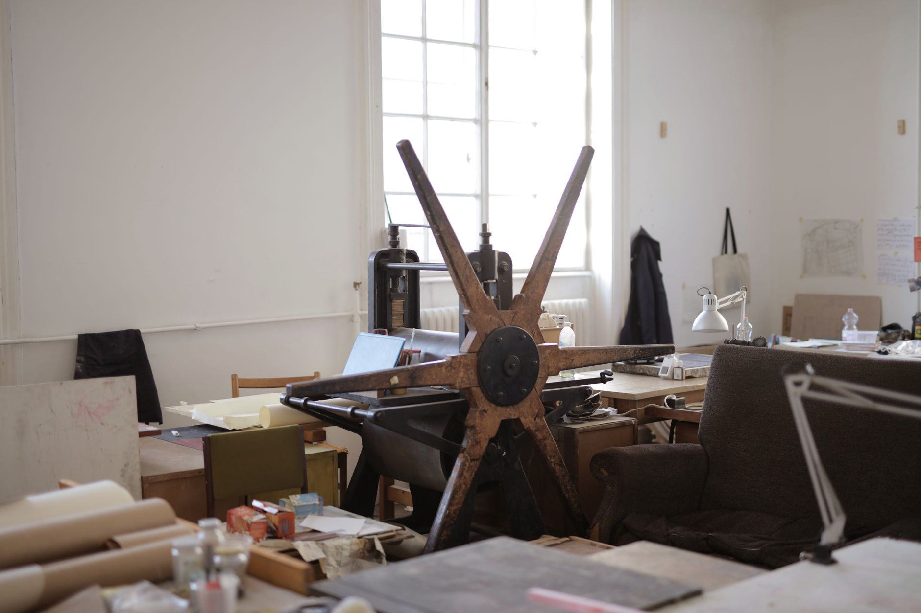 Atelier d’estampe avec une ancienne presse à étoile en bois au centre, entourée de tables, de rouleaux de papier, de flacons et d’une lampe.