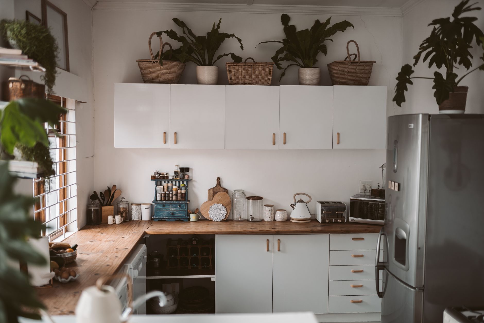 Cuisine aux meubles hauts blancs, paniers en osier et plantes posés sur le dessus, plan de travail en bois et réfrigérateur en inox.