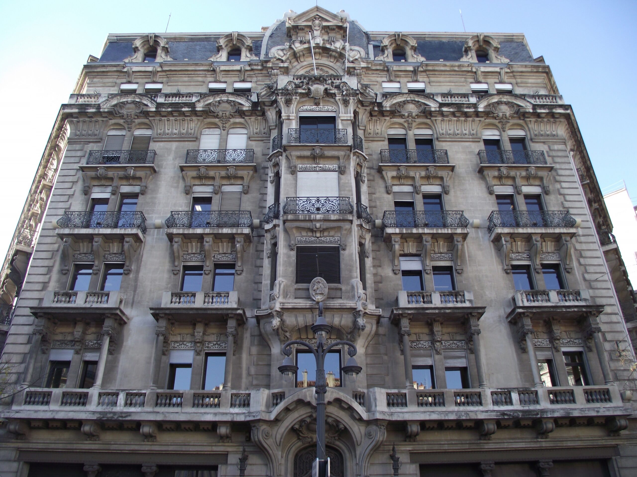 Façade en pierre ornée d’un immeuble ancien, avec balcons en fer forgé, balustrades et lampadaire, sous un ciel bleu.